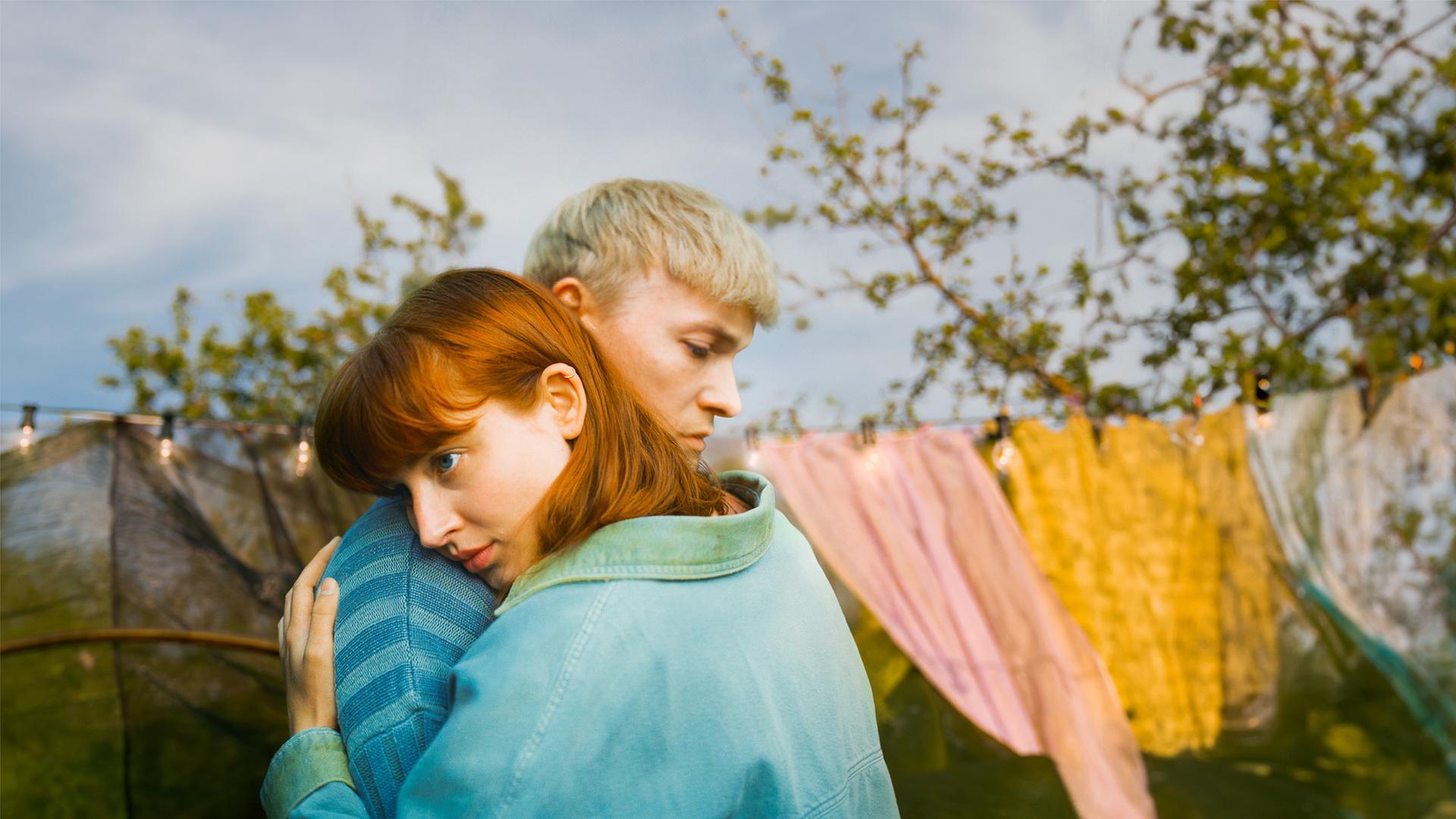 "Was du von mir sehen kannst": Gwen (Sina Genschel) trägt eine Jeansjacke mit grünem Kragen und grünen Jackenärmeln. Sie umarmt Adam, (Julius Nitschkoff) der einen blau-gestreiften Pullover trägt. Sie legt Ihren Kopf auf seine Schulter. Beide blicken traurig nach unten. Im Hintergrund sieht man eine Wäscheleine, auf der Wäsche trocknet.