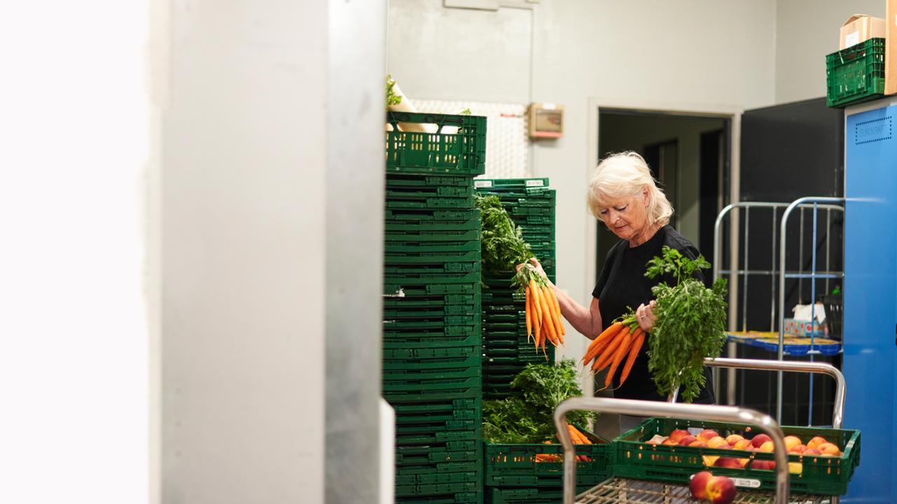 "37°: Rente? Reicht nicht!": Angelika steht in einem Lagerraum vor grünen, gestapelten Gemüsekisten. Sie hält zwei Bund Möhren in der Hand. Neben ihr steht ein Wagen mit einer Kiste mit Pfirsichen.