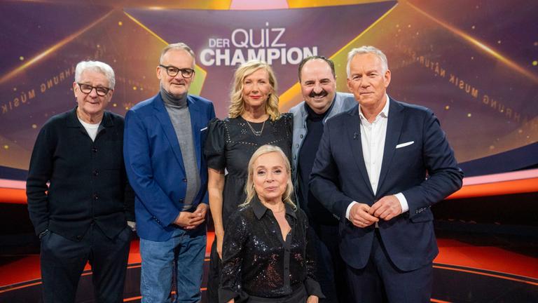 "Der Quiz-Champion": Gruppenfoto: Marcel Reif, Frank Plasberg, Andrea Kiewel, ChrisTine Urspruch, Johann Lafer und Johannes B. Kerner stehen vor dem Sendungslogo im Studio und lächeln in die Kamera.