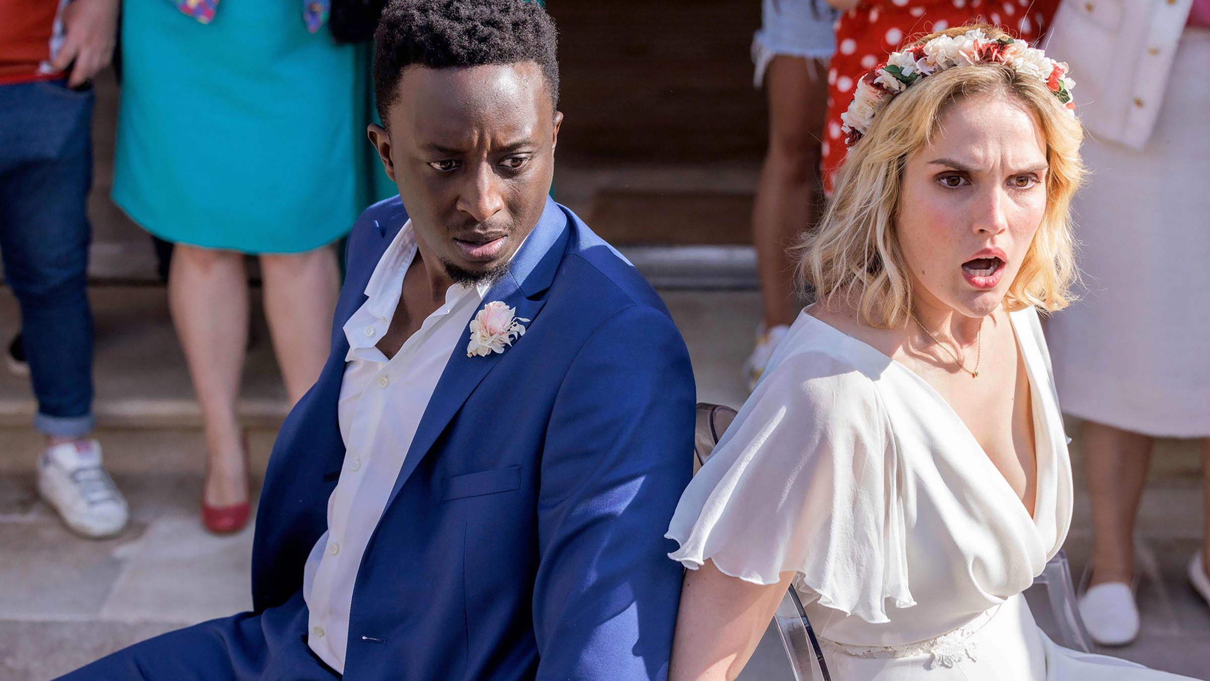 "Tiny Wedding - Unsere mega kleine Hochzeit": Halbnahe, leichte Aufsicht: Max (Ahmed Sylla) und Lou (Camille Lou) sitzen in ihren Hochzeitsoutfits mit schockierten Gesichtern Rücken an Rücken an Stühle gefesselt vor einer Feiergesellschaft. Diese ist nur hüftabwärts im Bild zu sehen.