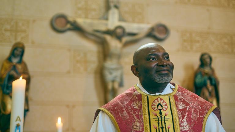 "Kirche ohne Rassismus?": Der nigerianische Prieser Uchenna Aba steht vor einem Kreuz und zwei Figuren und schaut lächelnd nach oben. Neben ihm brennt eine Kerze.