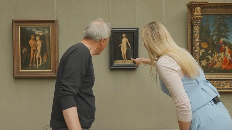 "Das Geheimnis der Meister: Lucas Cranach": Zwei Personen, ein Mann und eine Frau, stehen vor drei gerahmten Gemälden in einem Galerieraum. Die Frau zeigt auf das mittlere Bild, das eine nackte Frauenfigur vor einem schwarzen Hintergrund darstellt.