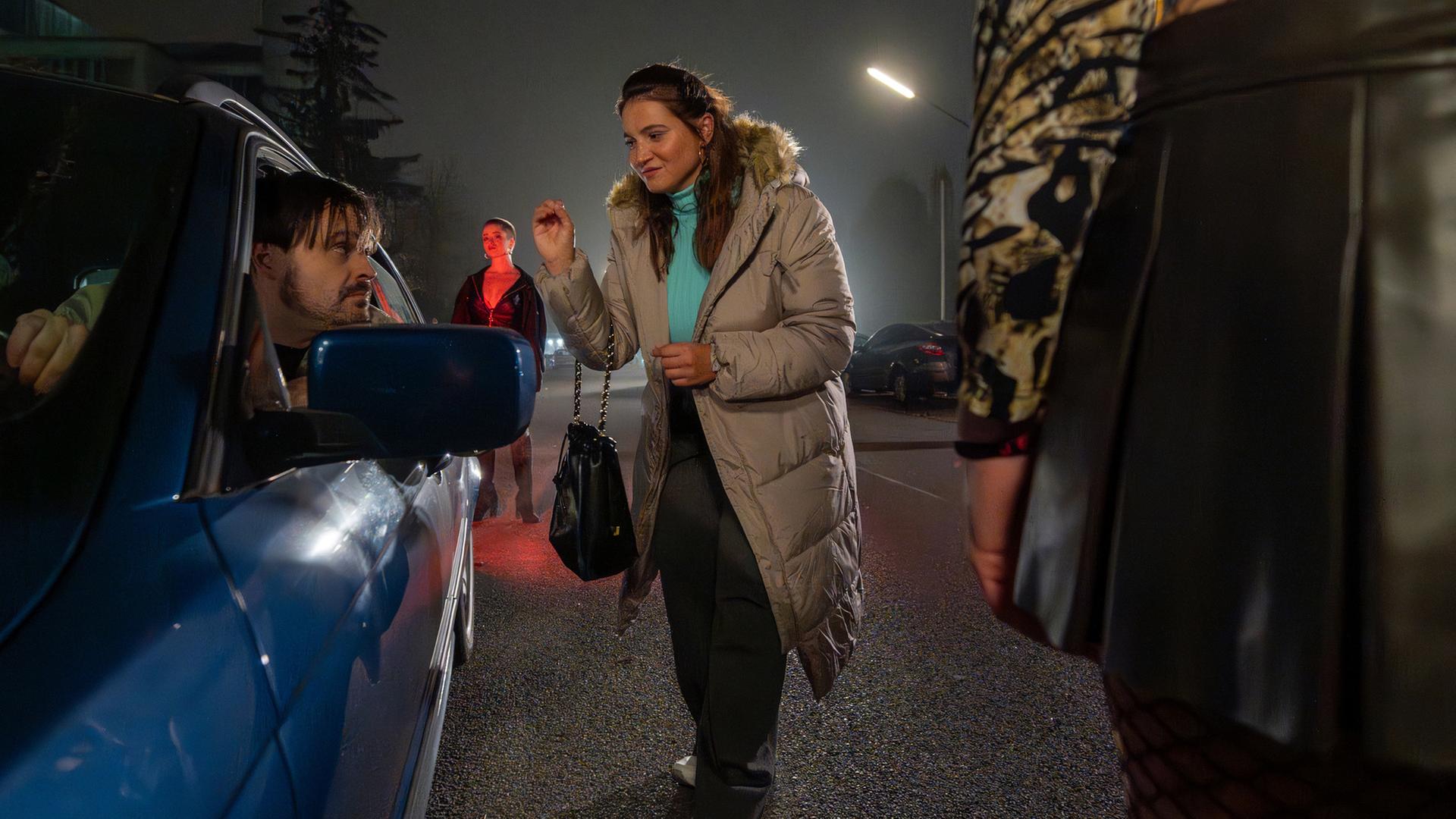 "Aktenzeichen XY... Cold Cases": Nachgestellte Szene Nachts auf einer Straße. Ein Auto steht am Straßenrand - der Fahrer schaut aus dem offenen Fenster und blickt zu einer jungen, hübschen Frau, die ihm mit der linken Hand ein Zeichen gibt. Beide schauen sich intensiv in die Augen. Im Hintergrund steht eine leicht bekleidete Frau und beobachtet das Geschehen.