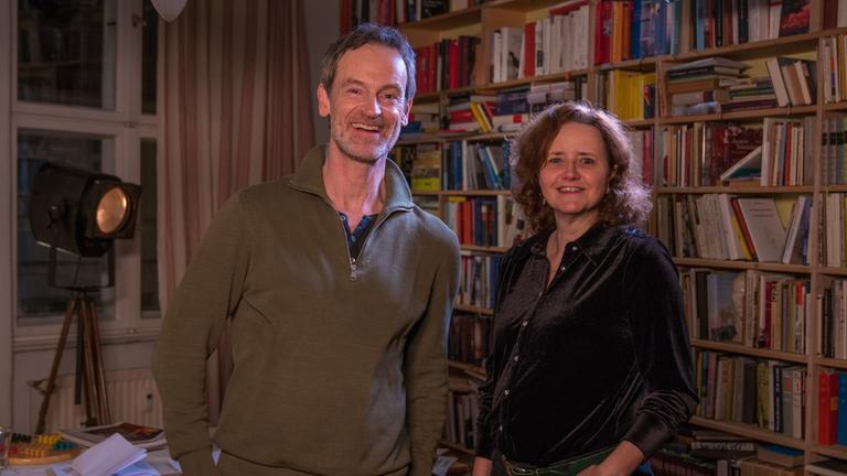 "Bitte um Rückruf - Interview mit einer Schriftstellerin": Schauspieler Jörg Hartmann und Schriftstellerin Julia Schoch stehen nebeneinander vor einer Bücherwand. Beide lächeln in die Kamera.