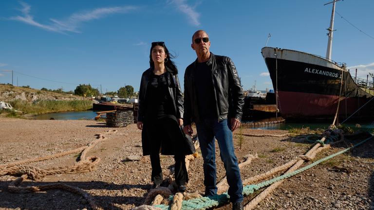 "Yvonne und der Tod": Ingo Thiel (Heino Ferch) und Lee Sooyoung (Kotbong Yang) stehen zwischen Tauen, die Schiffe im Hintergrund befestigen. Beide haben den Blick kritisch in die Ferne gerichtet. Ingo Thiel trägt eine Sonnenbrille.