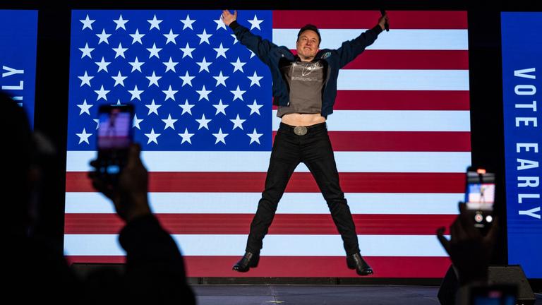 "Die Elon-Musk-Story - Superreich und supermächtig?": Elon Musk springt auf einer Bühne in die Luft, hinter ihm hängt die amerikanische Flagge.