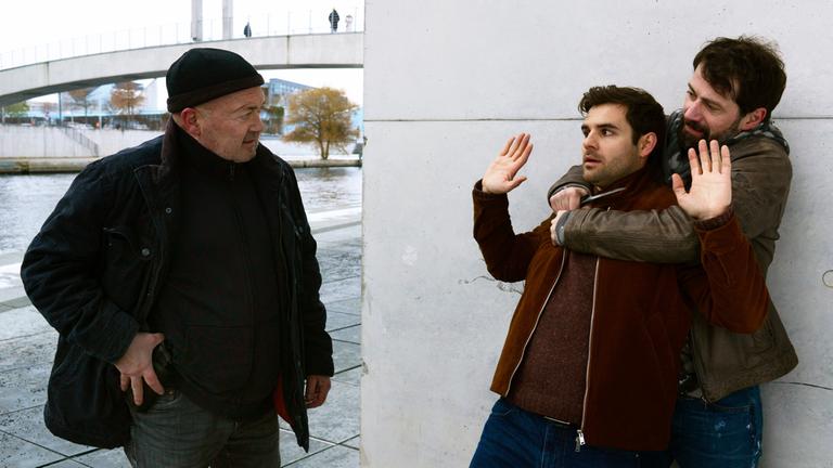 "Ein starkes Team - Fast perfekte Morde": Im Freien am Flussufer: Otto Garber (Florian Martens) blickt auf eine bedrohliche Situation und hält seine Hand an seine Dienstwaffe am Gürtel. Vor ihm hält Dennis Zander (Björn von der Wellen) den Ermittler Nils Makowski (Lucas Reiber) fest und hält ihm eine Messer an die Kehle.