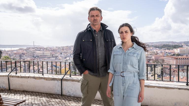 "Flucht aus Lissabon": Sophia (Hana Sofia Lopes) steht zusammen mit Tom Fährmann (Hans Sigl) auf einer bekannten Aussichtsplattform in Lissabon. Mit ernster und fragender Miene schauen beide in Richtung Kamera. Tom Fährmann hat die Hände in den Hosentaschen. Im Hintergrund sieht man die Häuser der Stadt von oben.