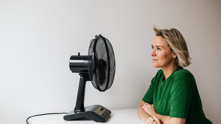 "Wechseljahre: heißkalt erwischt": Line sitzt vor einer weißen Wand an einem weißen Tisch. Auf dem Tisch steht ein schwarzer Tischventilator, auf den Line nachdenklich blickt.
