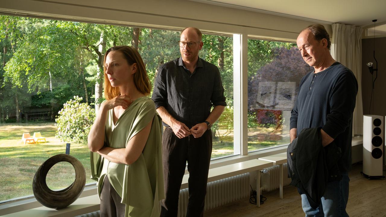 "Wendland - Stiller und der Teufelssauger": Jakob Stiller (Ulrich Noethen), Oda Nolde (Katharina Schüttler) und Jan Nolde (Johann von Bülow) stehen zusammen in einem Wohnzimmer. Oda Nolde steht mit dem Rücken zu Jakob Stiller und Jan Nolde. Hinter ihnen ist der Garten durch eine Fensterfront zu sehen.