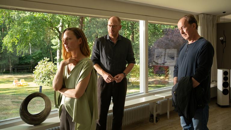 "Wendland - Stiller und der Teufelssauger": Jakob Stiller (Ulrich Noethen), Oda Nolde (Katharina Schüttler) und Jan Nolde (Johann von Bülow) stehen zusammen in einem Wohnzimmer. Oda Nolde steht mit dem Rücken zu Jakob Stiller und Jan Nolde. Hinter ihnen ist der Garten durch eine Fensterfront zu sehen.