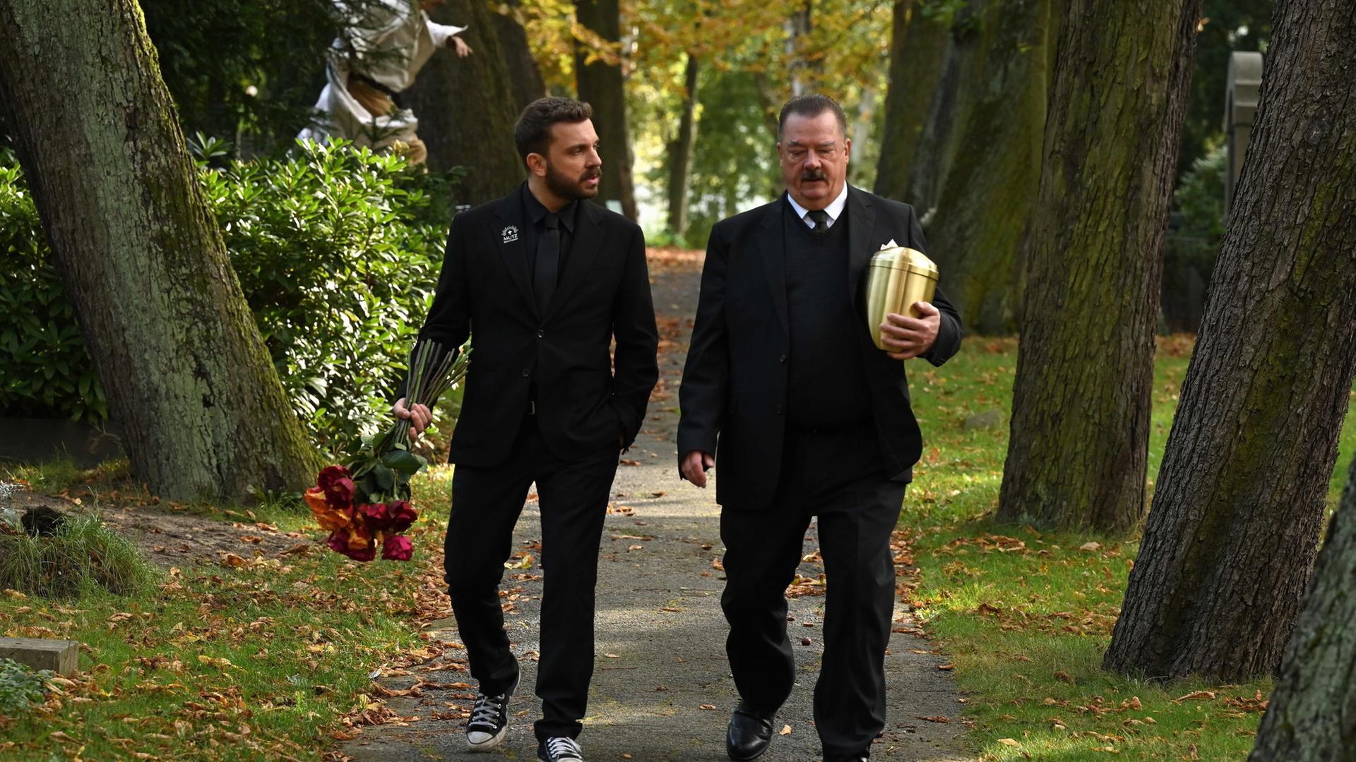 "Sterben für Beginner": Eric (Edin Hasanovic) und Volker Mutz (Peter Kurth) laufen mit Rosen und einer goldenen Urne über einen Friedhof mit alten Bäumen.