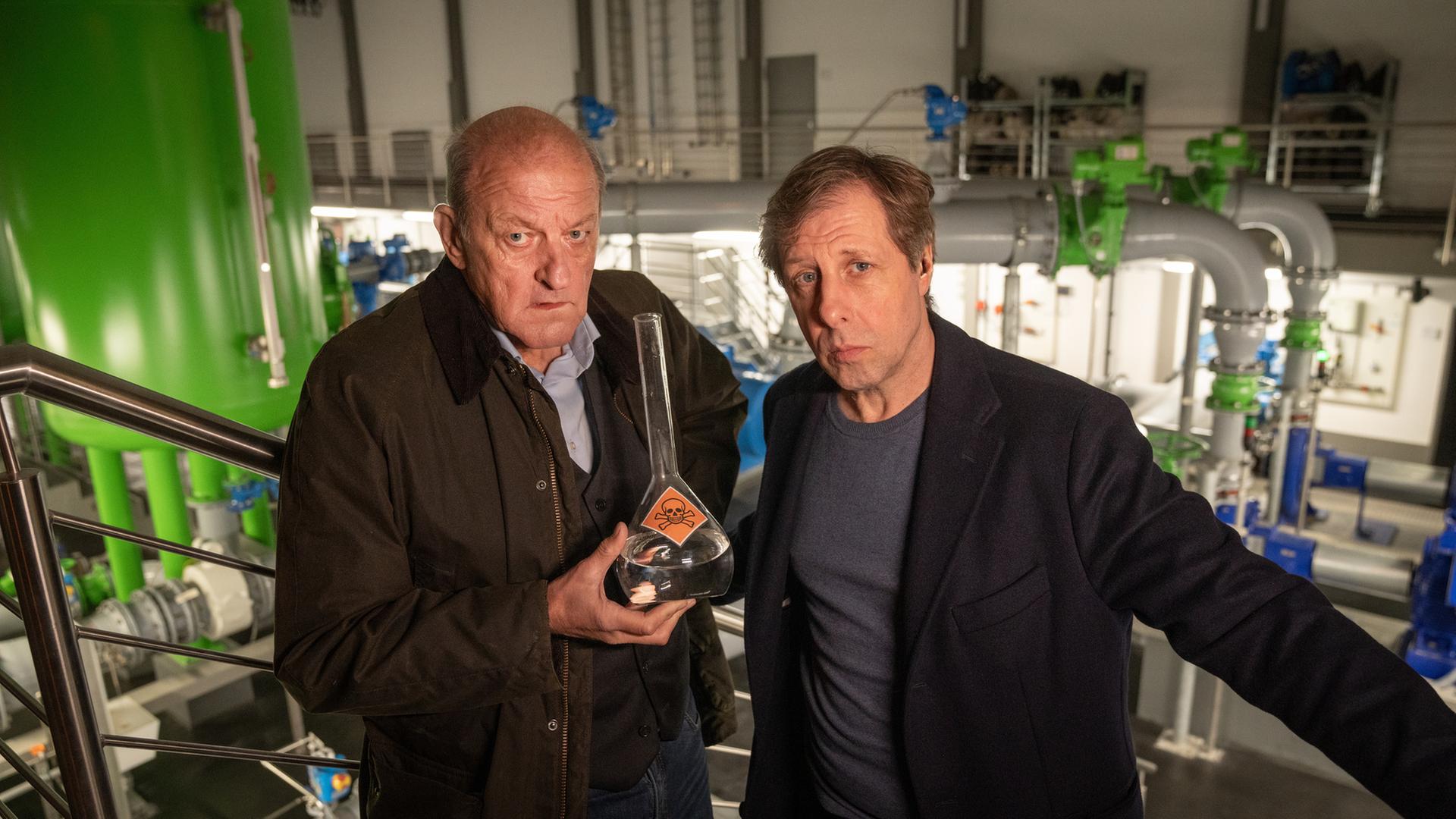"Wilsberg - Mit allen Wassern gewaschen": In einer Fabrikhalle stehen Wilsberg (Leonard Lansink) und Ekki (Oliver Korittke) nebeneinander und schauen ernst in die Kamera. Wilsberg hält einen Messkolben in der Hand, in welchem sich Wasser befindet und der mit einem Totenkopf-Warnaufkleber versehen ist.