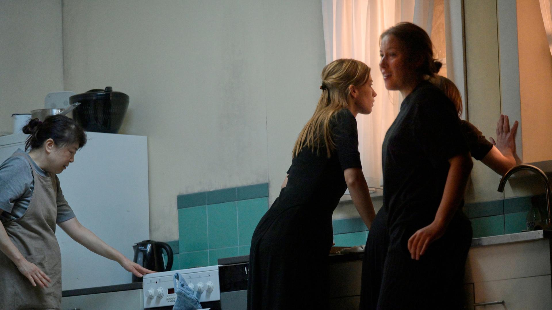 "Bernarda Albas Haus": Vier Frauen in einer Küchenkulisse. Links bedient eine Frau in Schürze den Herd. In der Mitte beugt sich eine Frau in schwarzer Kleidung über die Spüle und schaut angespannt nach draußen. Neben ihr steht eine weitere Frau in Schwarz, die sich leicht zur Seite dreht und mit einem Lächeln den Raum betrachtet. Die vierte Frau steht dicht am Fenster und legt eine Hand an die Scheibe, während ihr Blick nach draußen geht. Die Szene wirkt angespannt und geheimnisvoll, geprägt von kühlen Blau- und Grautönen.