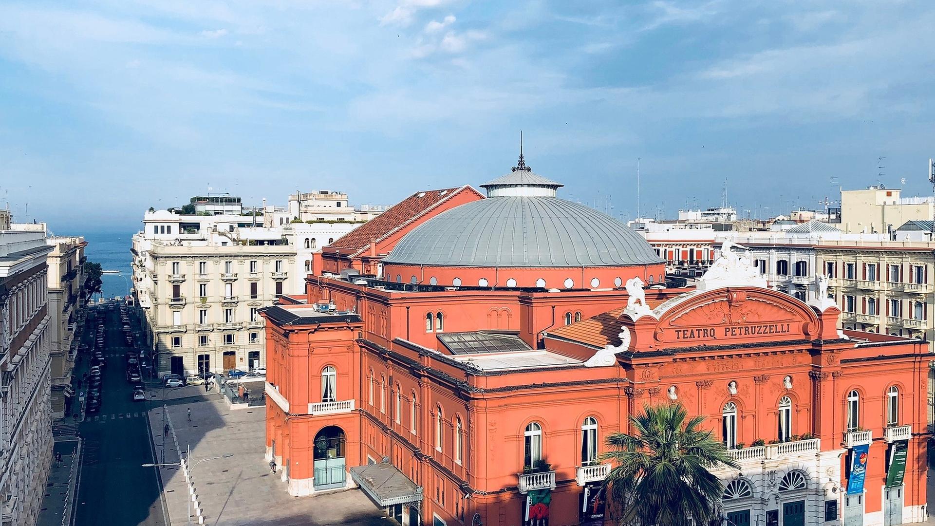 Europakonzert 2025 aus Bari mit den Berliner Philharmonikern und Riccardo Muti": Außenansicht des Teatro Petruzzelli im Stil des Neoklassizismus. Ein prachtvoller Bau aus rotem Stein und einem Kuppeldach mit weißen Fenstern und Türen liegt unter blauem Himmel.