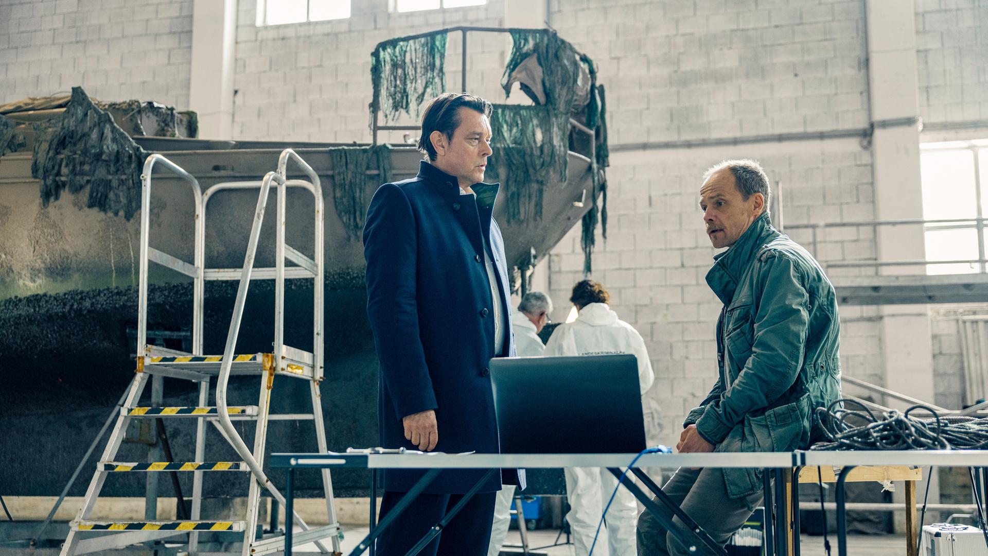 "Die Toten vom Bodensee - Das Geisterschiff": Thomas Komlatschek, (Hary Prinz) und Micha Oberländer (Matthias Koeberlin) stehen sich in einer Halle gegenüber. Hinter ihnen arbeiten Mitarbeiter der Spurensicherung (Komparsen) an einem Schiff.