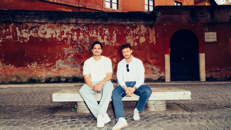 "Hummels - La Finale. Mit Tommi Schmitt": Mats Hummels und Tommi Schmitt sitzen auf einer Bank in Rom. Sie schauen in die Kamera.
