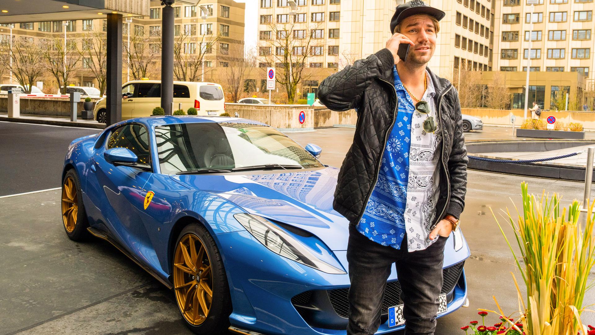 "Aktenzeichen XY... Vorsicht, Betrug!": Spielszene: Ein junger Mann steht, mit einem Smartphone telefonierend, lächelnd vor einem blauen Ferrari-Sportwagen mit goldenen Felgen. Im Hintergrund sind hohe Gebäude und etwas grün.