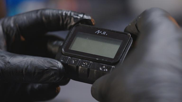 "frontal-Dokumentation: Operation Apollo - Die Pager Attacke des Mossad": Nachgestellte Szene, Nahaufnahme: Zwei Hände in Handschuhen halten einen Pager.