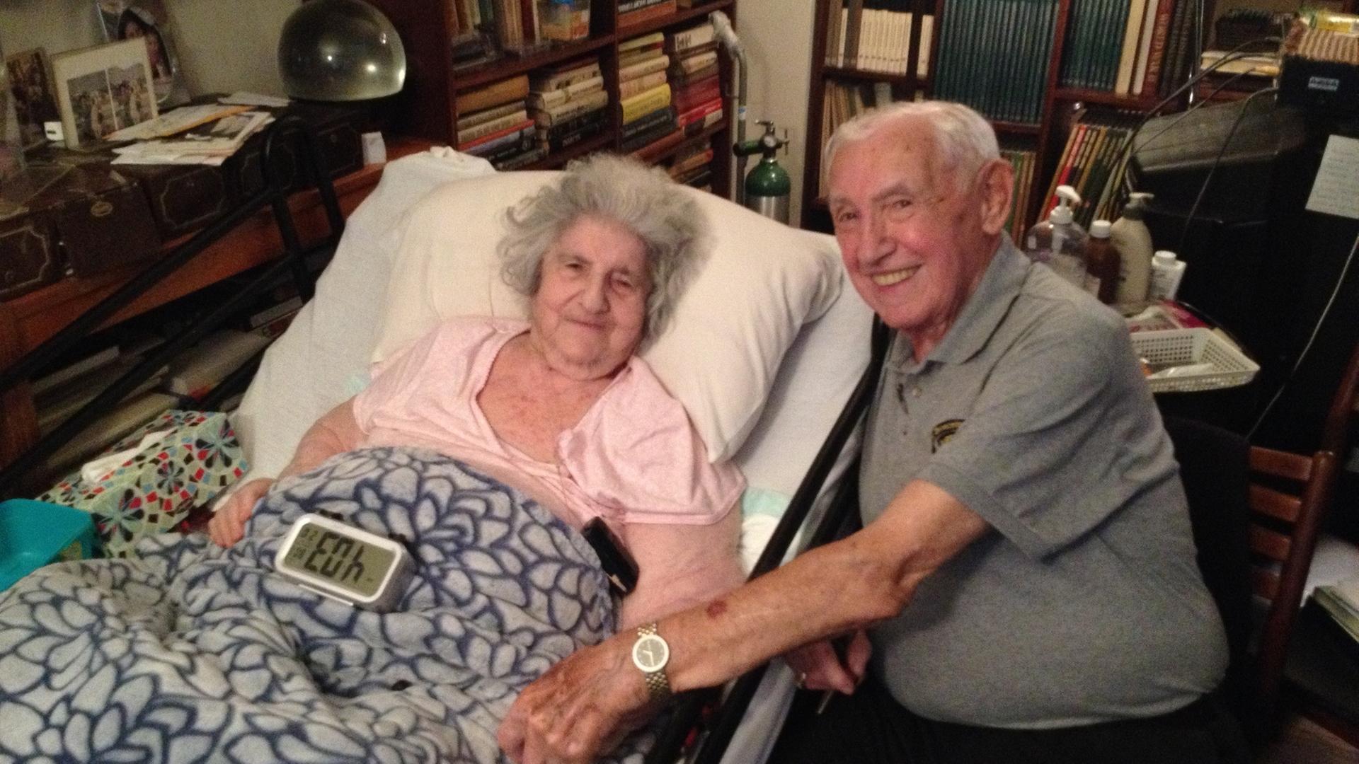 "Überleben in Auschwitz": Helen Spitzer, eine ältere Dame, liegt zu Hause in einem Krankenbett. David Wisnia, ein älterer Herr, sitzt an ihrem Bett und hält ihre Hand. Beide schauen in die Kamera.