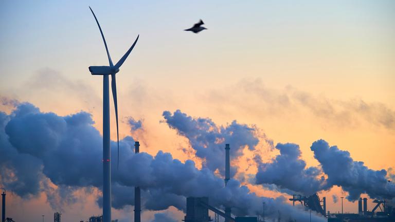 "planet e.: Durchbruch oder Scheitern - Wo steht der globale Klimaschutz?": Rauchschwaden aus Industrieschloten im Hintergrund vor orangegetöntem Abendhimmel. Im Vordergrund ein Windrad.