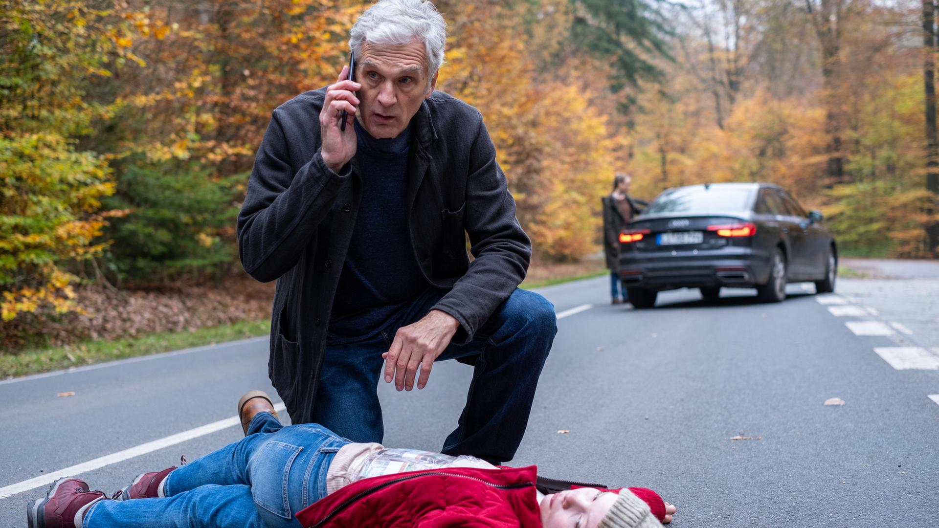 "Der Kommissar und der See – Das fremde Kind": Robert Anders (Walter Sittler) kniet hinter der am Boden liegenden Hannah Brunner (Laeni Geiseler) auf der Straße. Im Hintergrund steht ein Auto auf der Straße, eine Person steht in der Unschärfe an einer geöffneten Tür.