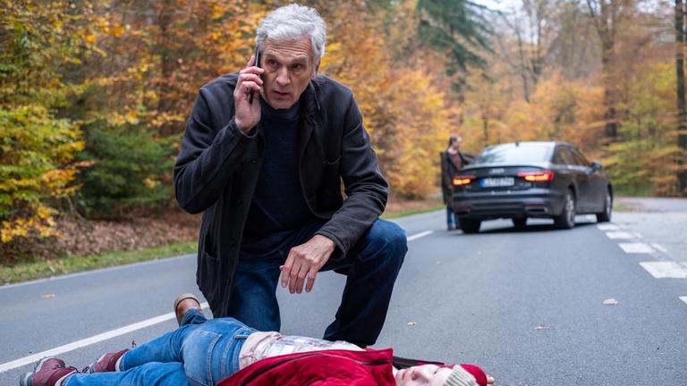 "Der Kommissar und der See – Das fremde Kind": Robert Anders (Walter Sittler) kniet hinter der am Boden liegenden Hannah Brunner (Laeni Geiseler) auf der Straße. Im Hintergrund steht ein Auto auf der Straße, eine Person steht in der Unschärfe an einer geöffneten Tür.