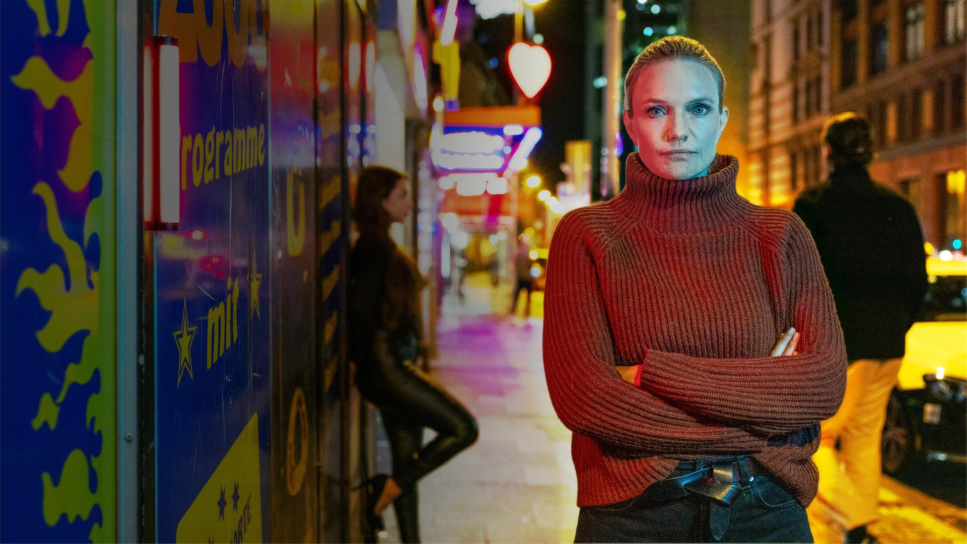 "Verbrechen! True Crime mit Sarah Tacke - Gefangen im Rotlicht": Sarah Tacke steht auf einem Gehweg und blickt in die Kamera. Im Hintergrund steht eine Prostituierte an eine Wand gelehnt. Neonreklamen tauchen die Straße in rötliches und gelbliches Licht.