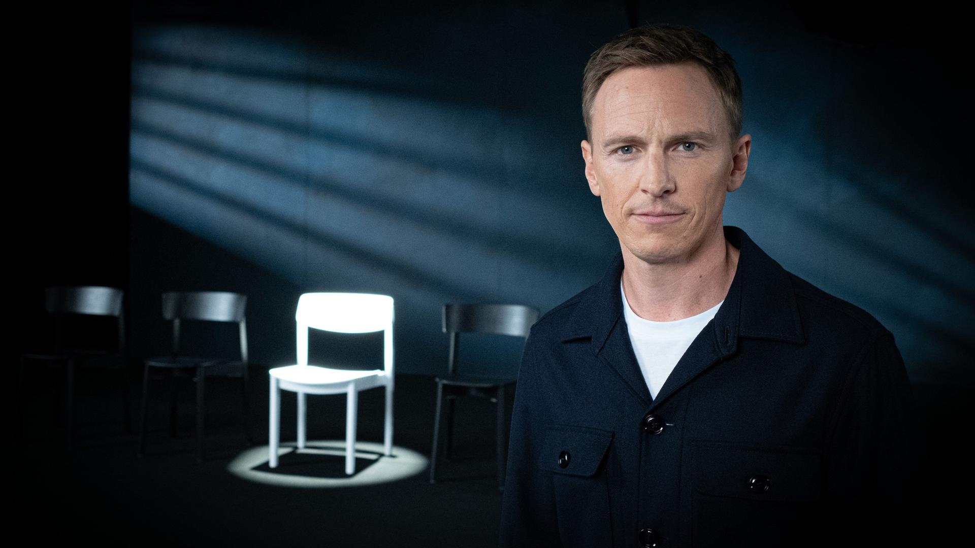 "Weil du mir gehörst! - Wenn Männer ihre Frauen töten": Jochen Breyer steht in einem abgedunkelten Raum und schaut in die Kamera. Im Hintergrund stehen mehrere Stühle, ein weißer Stuhl wird angestrahlt.