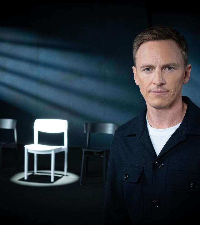 "Weil du mir gehörst! - Wenn Männer ihre Frauen töten": Jochen Breyer steht in einem abgedunkelten Raum und schaut in die Kamera. Im Hintergrund stehen mehrere Stühle, ein weißer Stuhl wird angestrahlt.