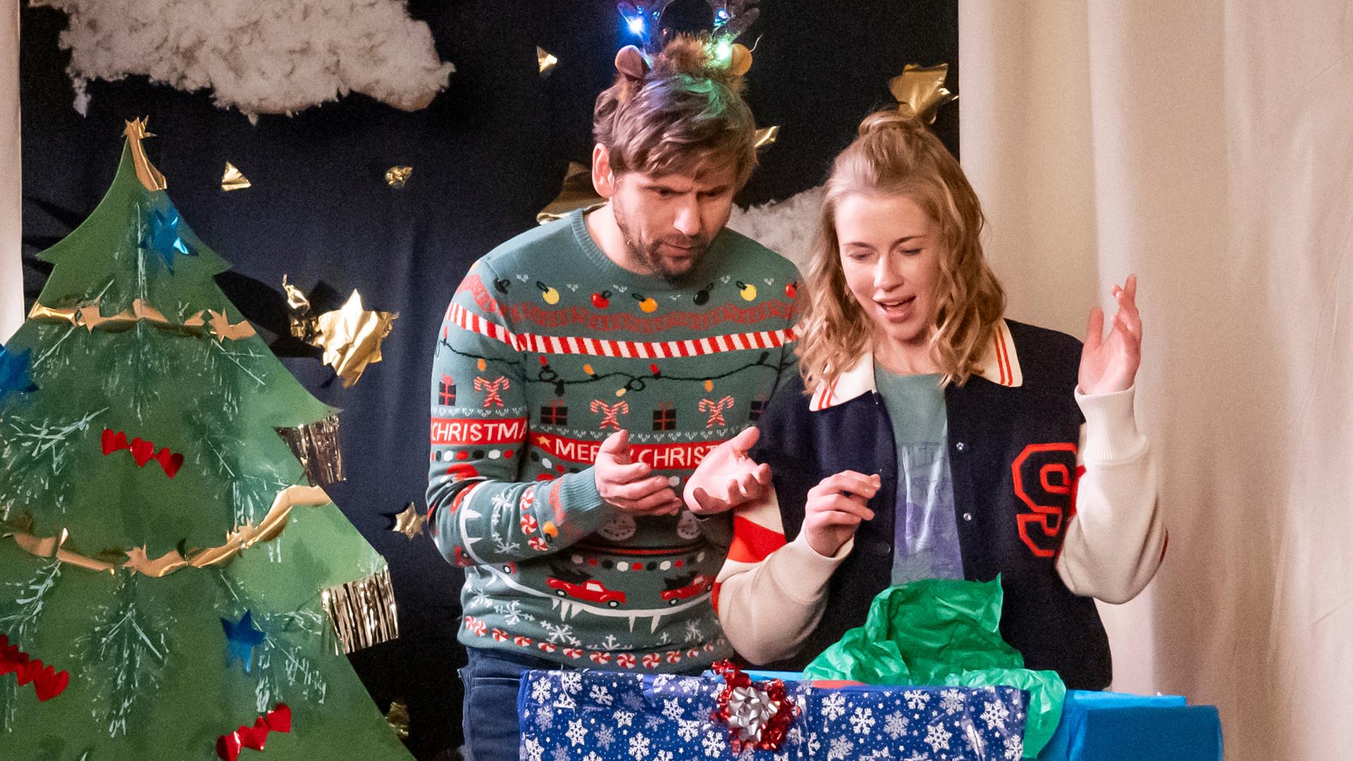 "Eine fast perfekte Bescherung": Bodo Hartmann (Steve Windolf) und Liz Schott (Christine Eixenberger) stehen auf einer Schulbühne hinter einem überdimensional großen Geschenkpaket und betrachten den Inhalt überrascht und amüsiert. Links hinter ihnen befindet sich ein geschmückter Weihnachtsbaum aus Pappe.