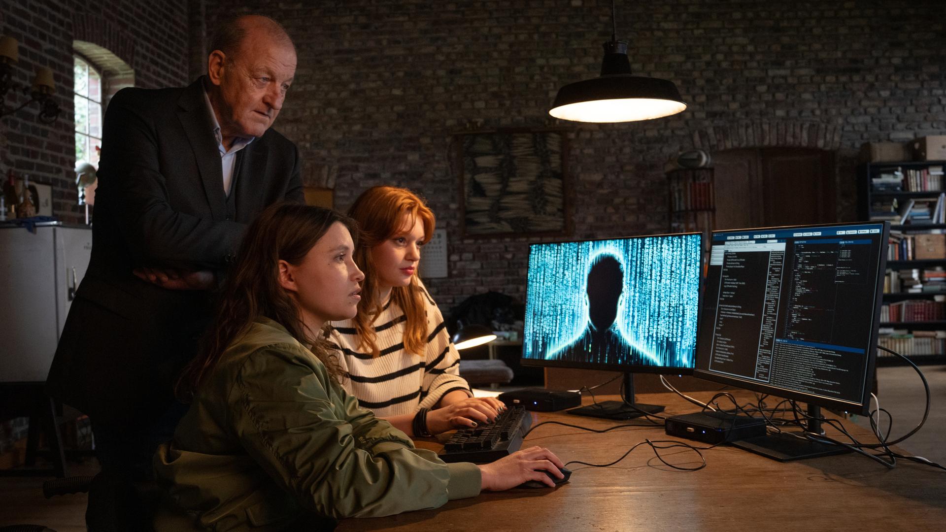 "Wilsberg - Phantomtod": Jana Haumann (Lilly Charlotte Dreesen) und Merle (Janina Fautz) sitzen an einem Tisch und bedienen eine Computer-Maus und eine Tastatur, während sie gebannt auf die beiden Bildschirme vor sich starren. Wilsberg (Leonard Lansink) steht hinter ihnen und blickt ebenfalls auf die Monitore, auf denen unter anderem eine dunkle Silhouette einer Person zu sehen ist.