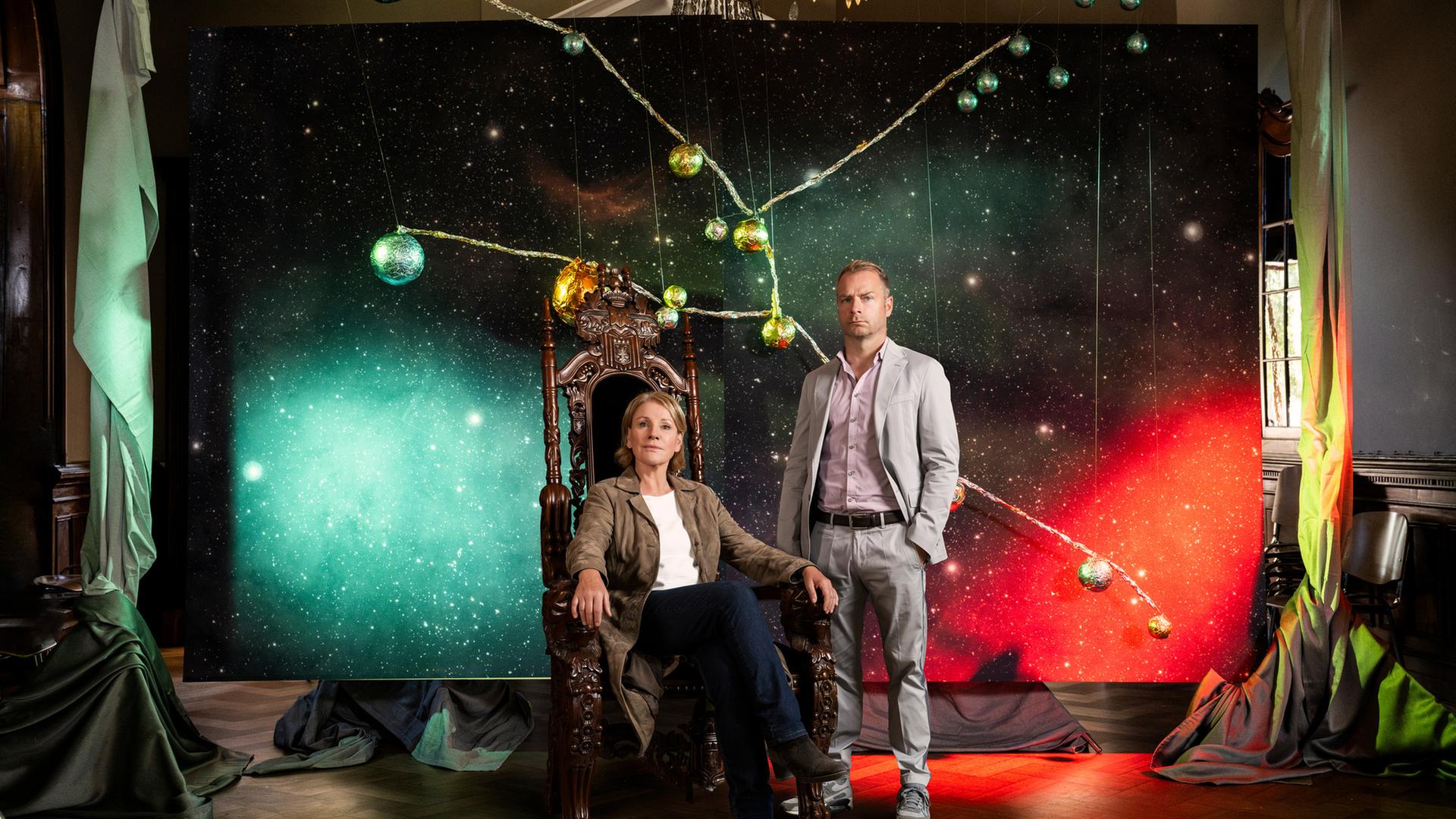 "Marie Brand und die Bedrohung vom anderen Stern": Marie Brand (Mariele Millowitsch) und Jürgen Simmel (Hinnerk Schönemann) posieren vor einem Sternenbild und schauen in die Kamera. Marie Brand sitzt dabei in einem alten prunkvollen Sessel.