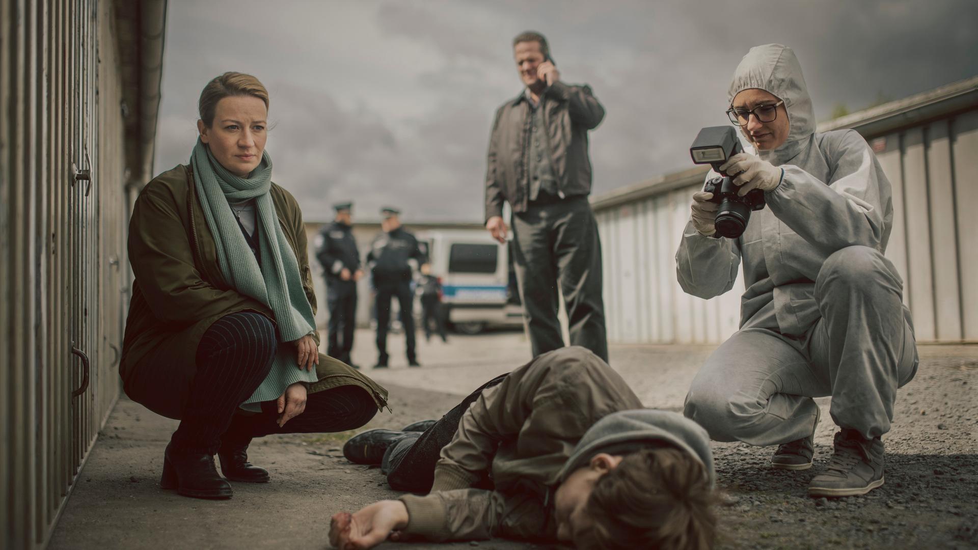 "Theresa Wolff - Nebel": Auf einem Garagenhof liegt Finn Behrend (Philip Günsch) tot, seitlich auf dem Boden. Dr. Theresa Wolff (Nina Gummich) kniet daneben während eine Mitarbeiterin der Spurensicherung (Komparsin) die Leiche fotografisch dokumentiert. Im Hintergrund telefoniert Bruno Lewandowski (Aurel Manthei) mit seinem Handy.