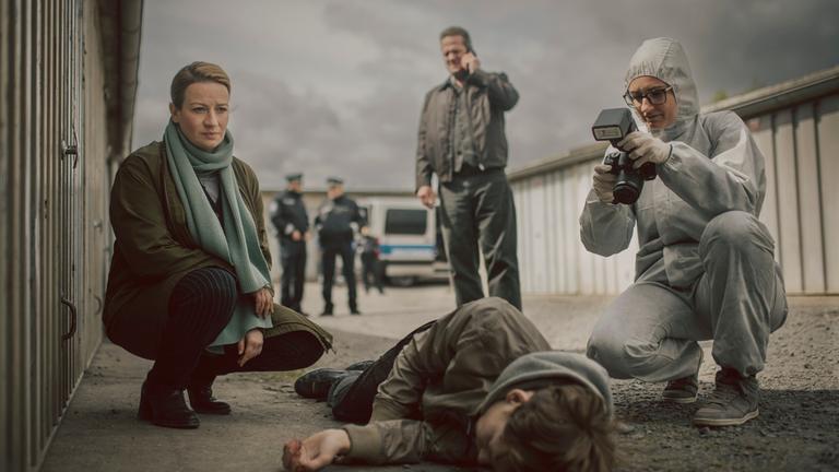 "Theresa Wolff - Nebel": Auf einem Garagenhof liegt Finn Behrend (Philip Günsch) tot, seitlich auf dem Boden. Dr. Theresa Wolff (Nina Gummich) kniet daneben während eine Mitarbeiterin der Spurensicherung (Komparsin) die Leiche fotografisch dokumentiert. Im Hintergrund telefoniert Bruno Lewandowski (Aurel Manthei) mit seinem Handy.