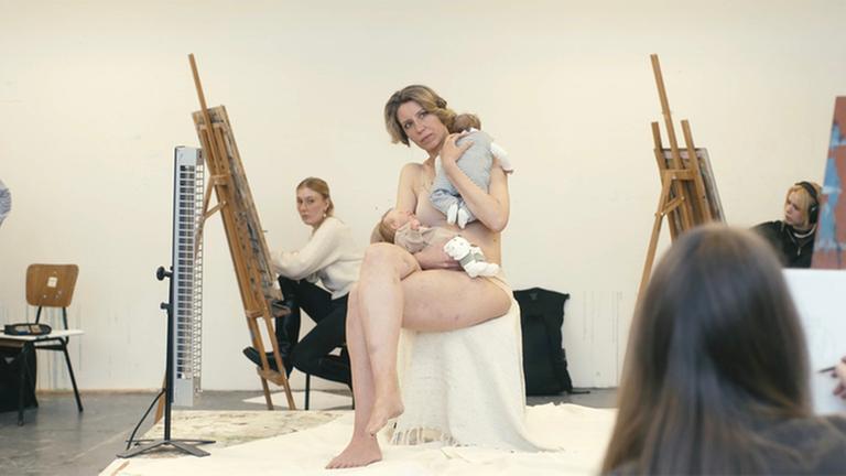 "Reproduktion": Eine Frau in Unterwäsche sitzt mit übereinandergeschlagenen Beinen in einem Atelier auf einem Hocker. In der rechten Armbeuge hält sie einen Säugling, mit dem linken Arm einen weiteren Säugling an ihrer linken Schulter. Um sie heruium vier Maler:innen an ihren Staffeleien. 