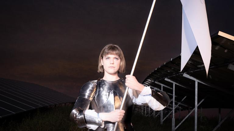 "Sonnenkönigin" (3sat): Johanna (Llewellyn Reichman) steht nachts vor einer Brücke. Sie trägt eine Ritterrüstung und schwenkt eine weiße Fahne.