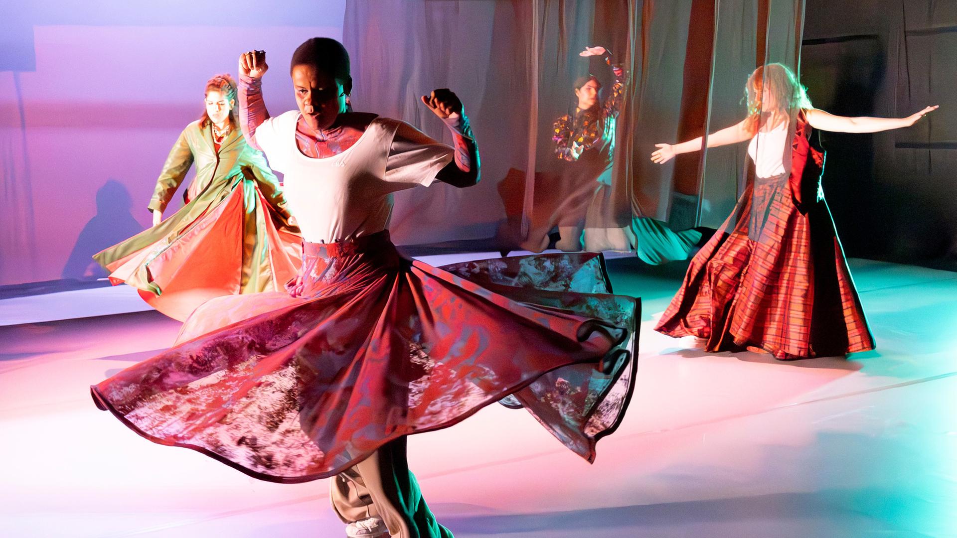 "Der Wert der Kultur – Der Deutsche Theaterpreis DER FAUST": Tanzszene auf einer hell ausgeleuchteten Bühne. Mehrere Personen tragen weite, farbenprächtige Kostüme in Rot-, Grün- und Violetttönen. Im Vordergrund dreht sich eine Person mit ausladendem Rock. Im Hintergrund sind weitere Tänzerinnen in dynamischen Posen zu sehen, transparente Stoffbahnen hängen als Bühnenbild von der Decke.