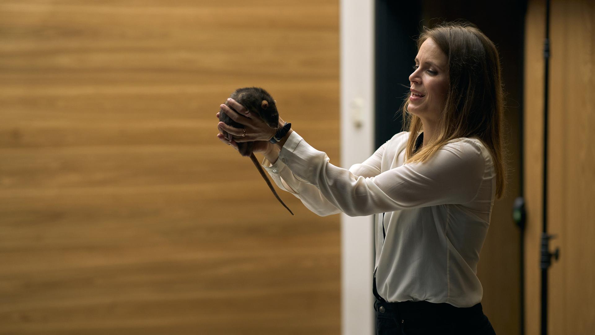 "Elixir – The Pain Killers - Aspergon": Isabelle (Hanna Verboom) steht in einem Konferenzraum vor einer holzgetäfelten Wand und einer Tür. Sie hält mit beiden Händen eine Ratte in die Höhe.