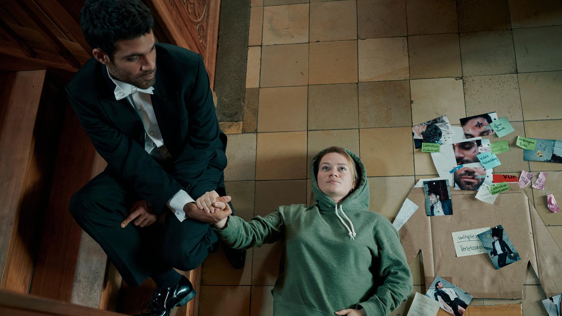 "Theresa Wolff - Passion": Blick von schräg oben: Gábor Kértesz (Deniz Arora) sitzt auf einer Holztreppe und hält die Hand von Dr. Theresa Wolff (Nina Gummich), die neben einem improvisierten Mordfallbrett auf dem Boden liegt.