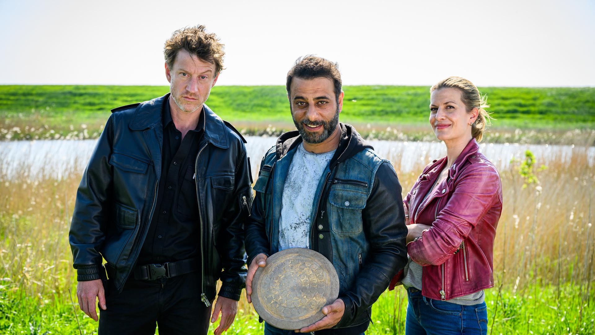 "Friesland - Tief im Dreck": Insa Scherzinger (Theresa Underberg), Yunus Özlügül (Yunus Cumartpay) und Henk Cassens (Maxim Mehmet) stehen vor einem Gewässer und einem grünen Deich, wobei Yunus eine Bronzescheibe in den Händen hält und lächelt.