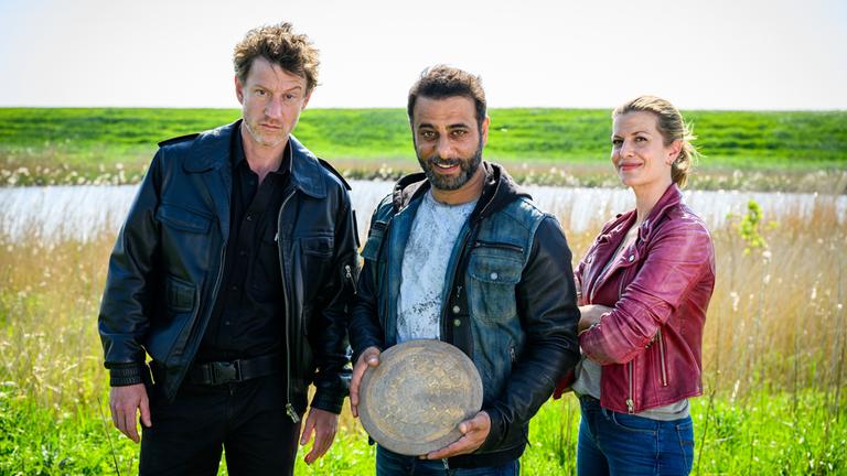 "Friesland - Tief im Dreck": Insa Scherzinger (Theresa Underberg), Yunus Özlügül (Yunus Cumartpay) und Henk Cassens (Maxim Mehmet) stehen vor einem Gewässer und einem grünen Deich, wobei Yunus eine Bronzescheibe in den Händen hält und lächelt.