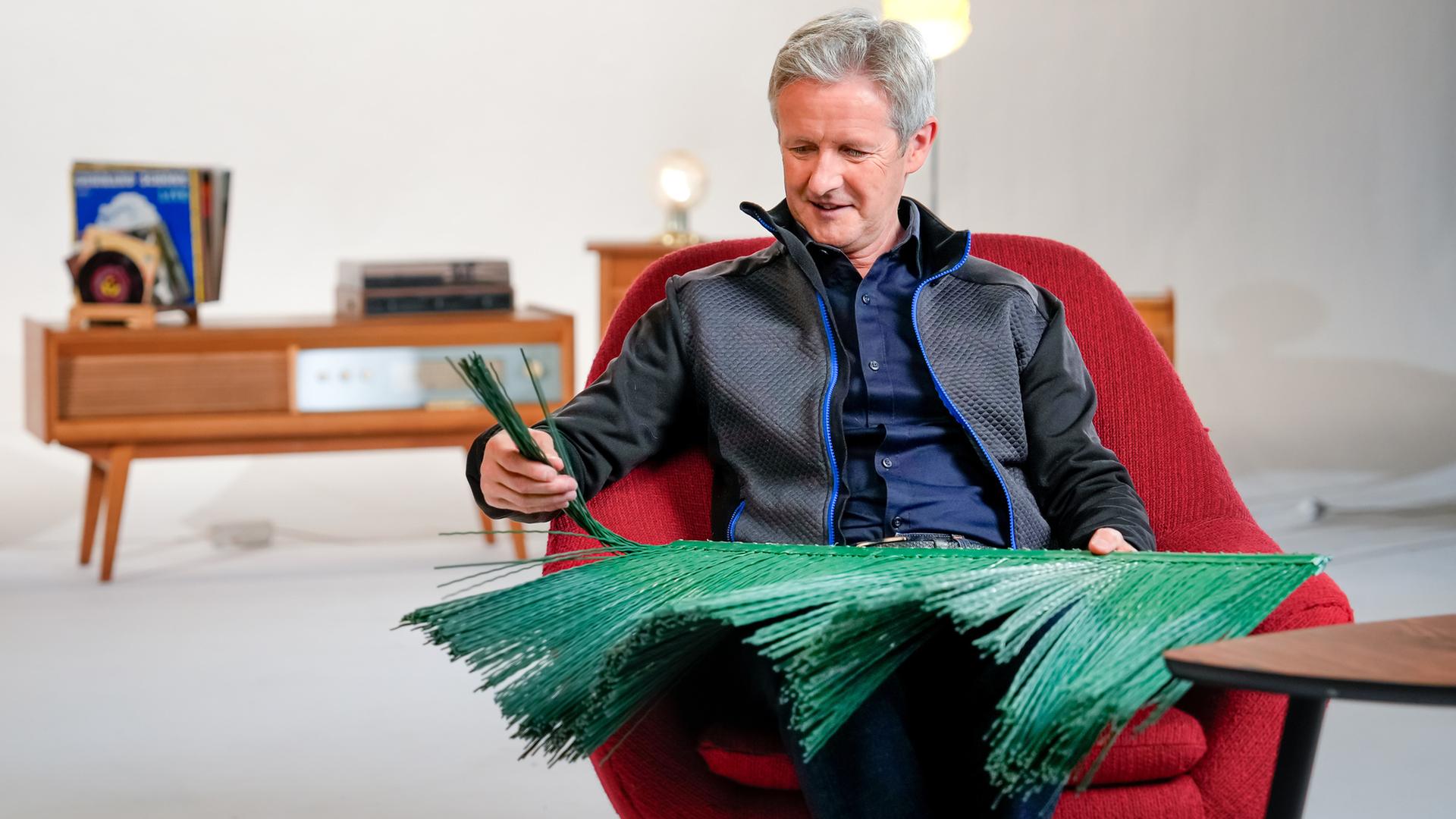 "DDR genial - Mangel macht erfinderisch": Jens Weißflog sitzt in einem roten Sessel, auf dem Schoß hält er ein großes bürstenartiges Element aus grünem Kunststoff. Seine Hand hält ein paar Bürstenstränge hoch. Im Hintergrund stehen Retro-Möbel.