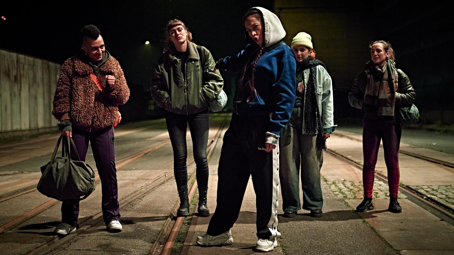 "Stralsund – Ablaufdatum": Der "Female Fight Club" steht bei Nacht auf einem Schienengelände. Gyzem (Zandile Amy Ngono), Jule (Sophie Pfennigstorf), Martha (Lo Rivera), Nina (Cheyenne Demont) und Hanna (Ramona Schlenker) stehen nebeneinander und schauen Richtung Kamera.
