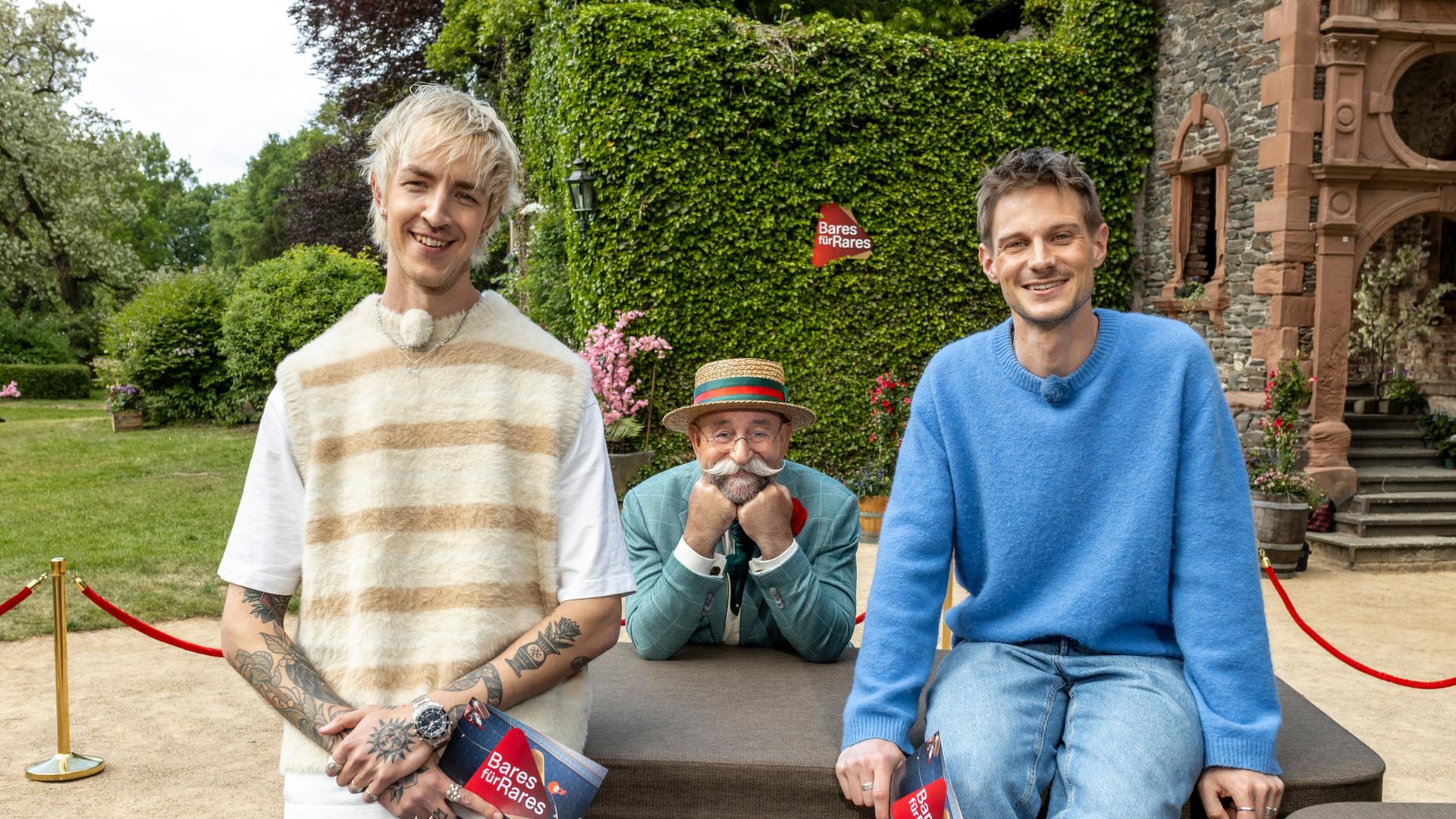 "Bares für Rares XXL - Voll im Trend": Vor einem Gebäude von Gut Nettehammer: Niklas van Lipzig steht vor dem Expertisenpult, David Martin sitzt darauf. Zwischen ihnen ist Horst Lichter zu sehen, der sich zum Pult beugt, sein Gesicht in beide Hände gestützt. Alle drei lächeln in die Kamera.
