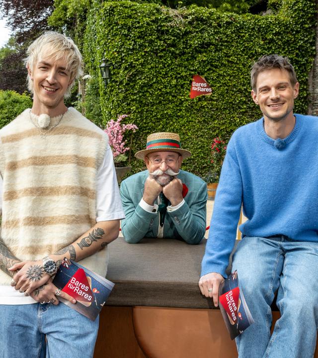 "Bares für Rares XXL - Voll im Trend": Vor einem Gebäude von Gut Nettehammer: Niklas van Lipzig steht vor dem Expertisenpult, David Martin sitzt darauf. Zwischen ihnen ist Horst Lichter zu sehen, der sich zum Pult beugt, sein Gesicht in beide Hände gestützt. Alle drei lächeln in die Kamera.