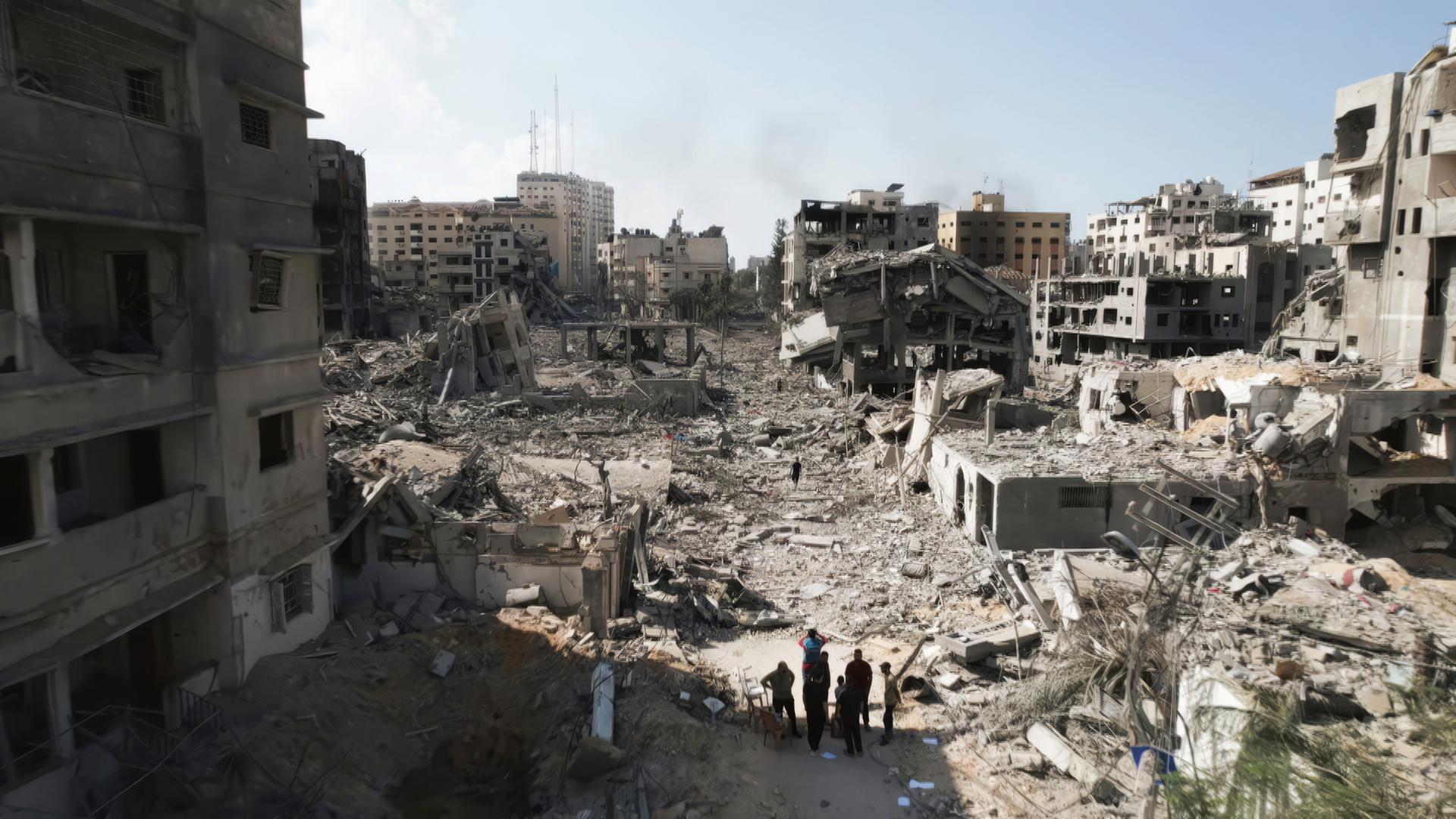  "Netanjahu, die USA und der Krieg in Gaza": Inmitten eingestürzter Gebäude nach Raketenangriff steht eine kleine Gruppe von Menschen. Teilweise steigt schwarzer Rauch von zerstörten Gebäuden auf.