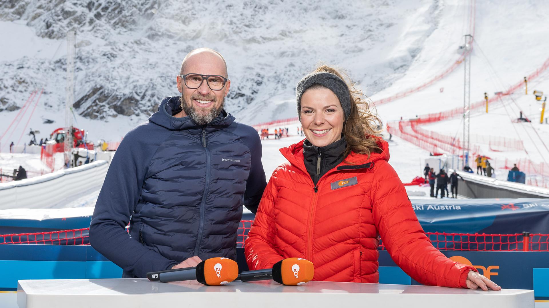  ZDF-Experte Ski, Marco Büchel, und ZDF-Moderatorin Ski Alpin, Amelie Stiefvatter, stehen am Presenter-Tisch im Freien vor der Skistrecke.