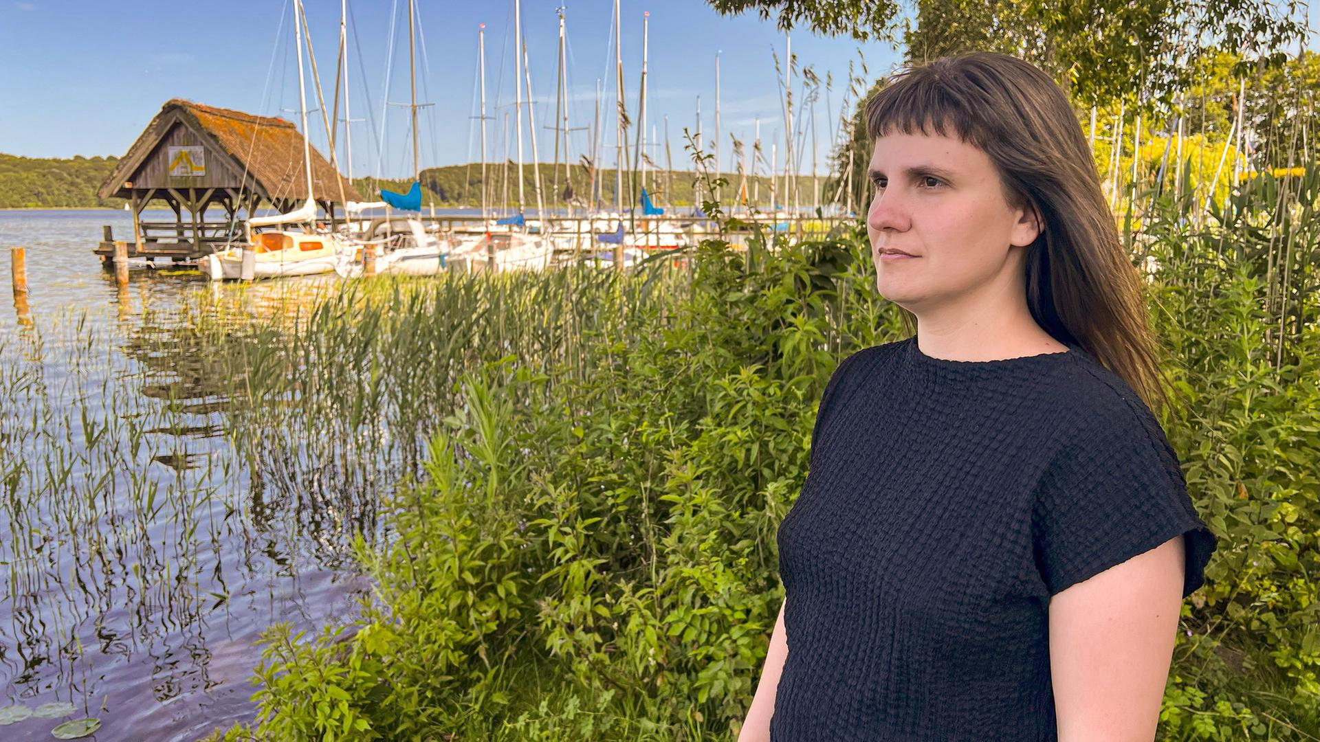 "Tatort Land: Rettung aus häuslicher Gewalt": Miriam steht am See und schaut in die Ferne. Hinter ihr ist die Böschung, dahinter ein Steg mit Segelbooten und ein Bootshaus.