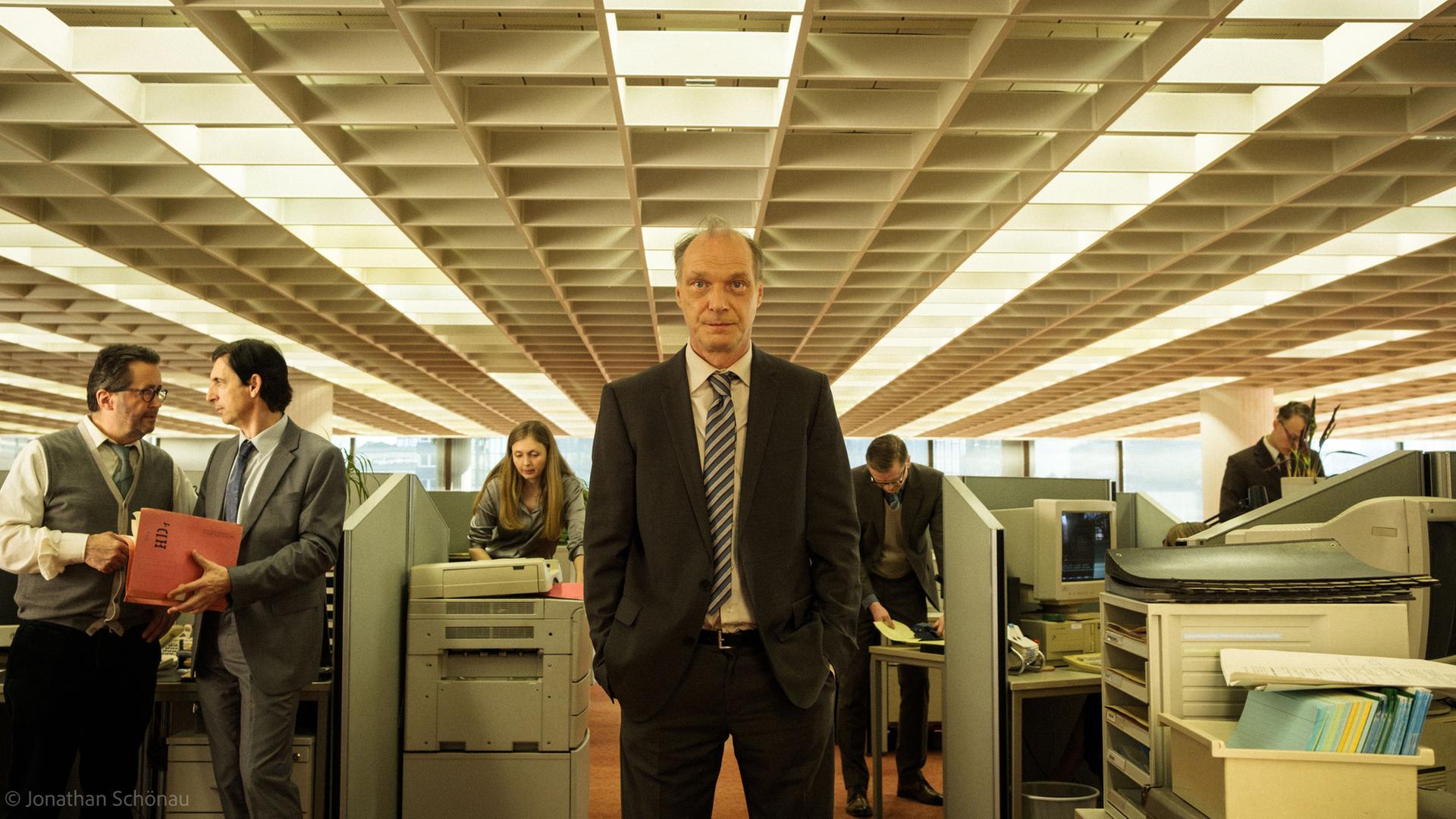 "Die Wahrheit über Arbeit und Geld - Was verdient Deutschland?": Spielszene: Versicherungskaufmann Schrader (Martin Brambach) steht in einem Großraumbüro und schaut nach vorne. Im Hintergrund befinden sich weitere Personen (Komparsen) zwischen Schreibtischen und einem Kopirer.